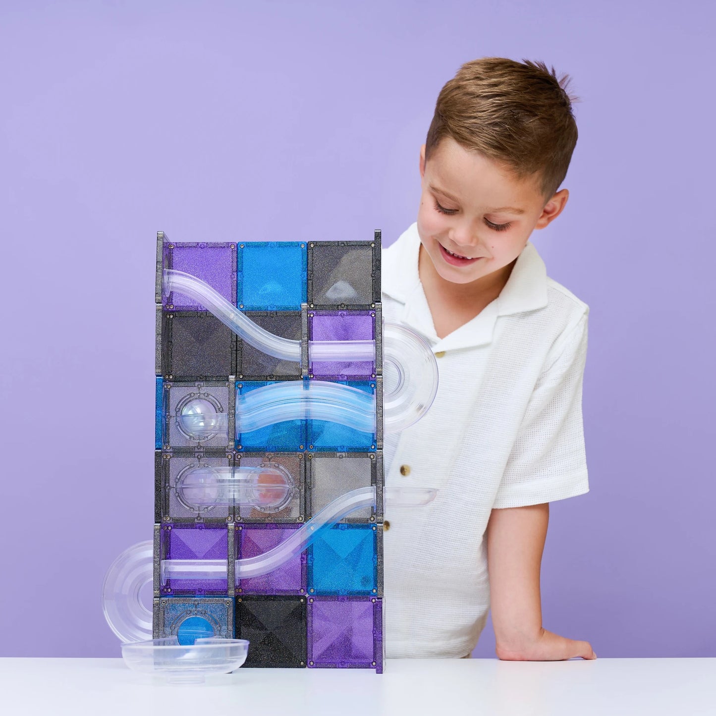 A young boy in a white polo shirt smiles while looking at a magnetic marble run toy
