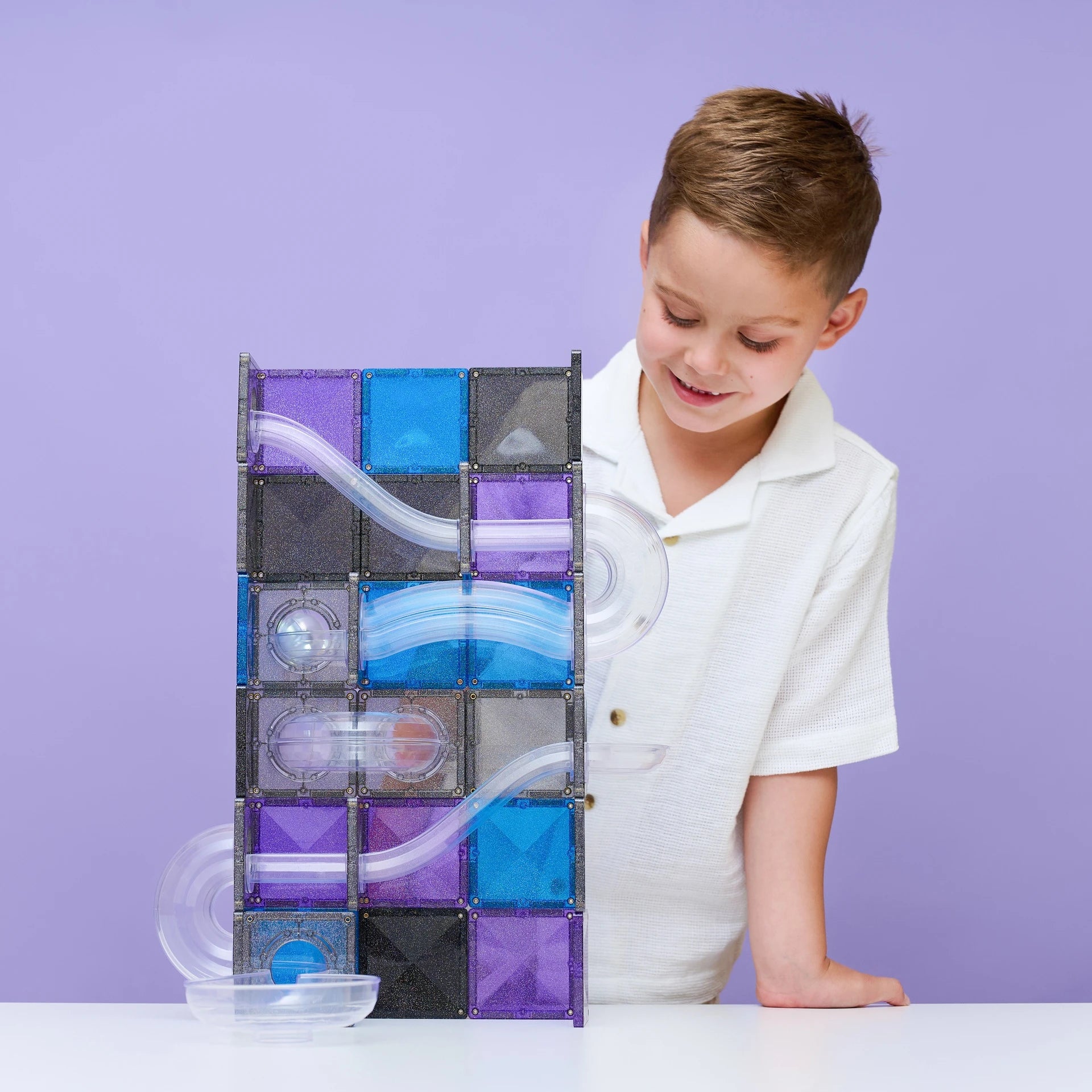 A young boy in a white polo shirt smiles while looking at a magnetic marble run toy