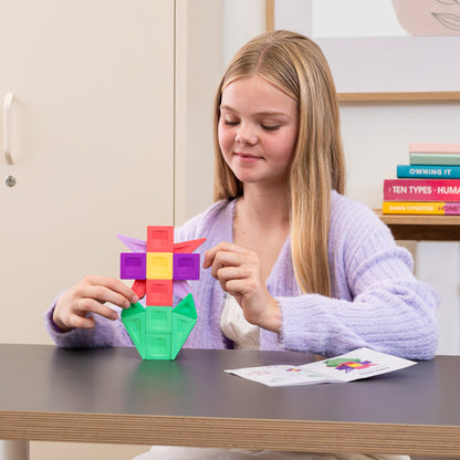 A young girl with blonde hair sits at a wooden desk, assembling a colorful plastic building to