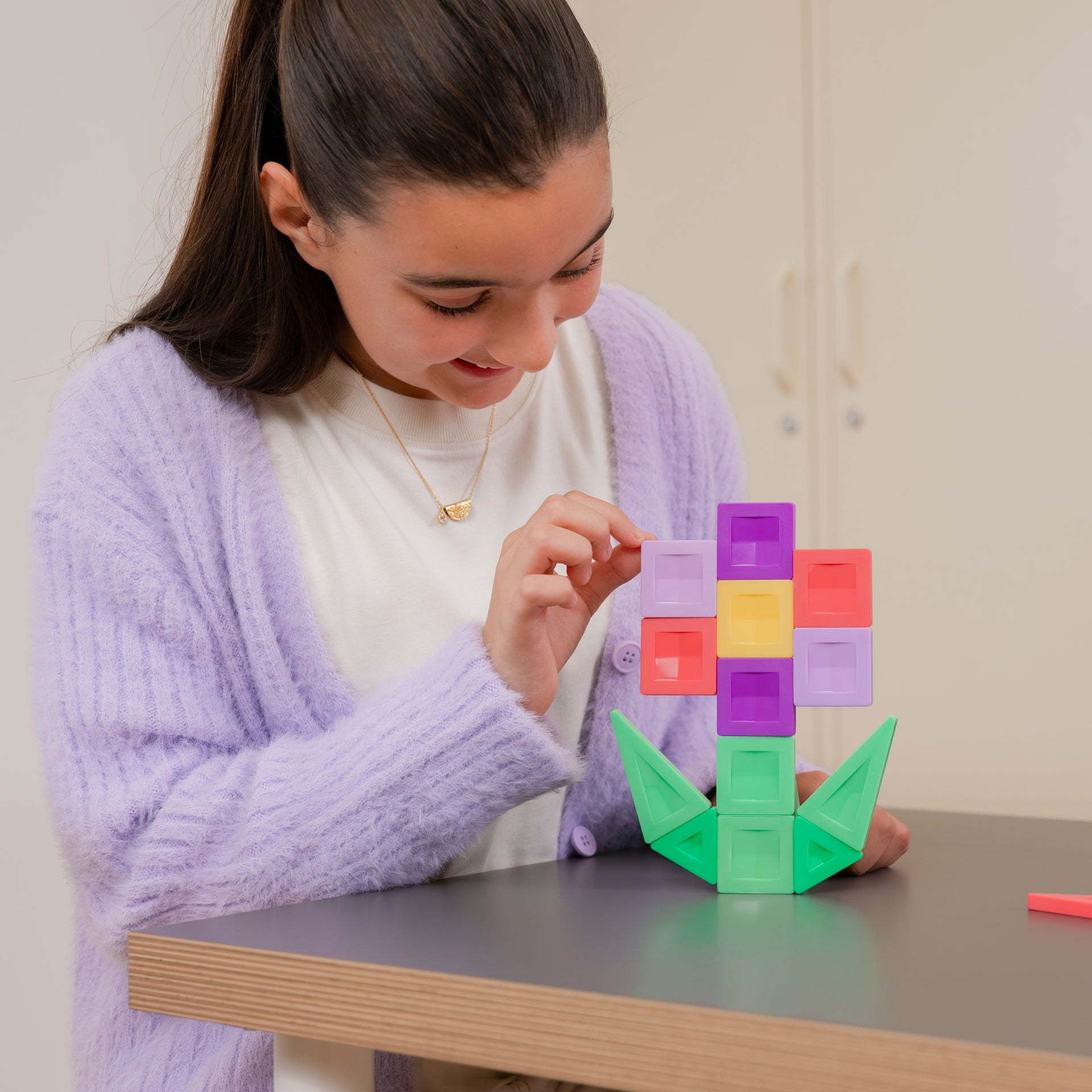 A young woman with dark hair in a ponytail is smiling while building a colorful toy structure with plastic blocks